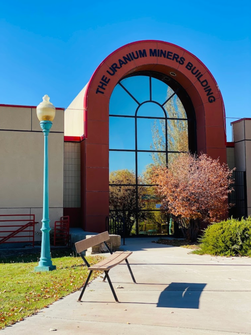 A bench in front of the Uranium Miners Building, featuring a large arched entrance and clear blue sky.